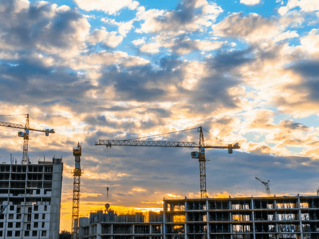 Image of a skyline containing a construction sites with cranes and a cloudy sky.